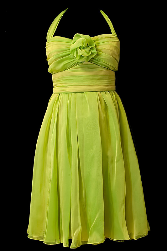 Krótka zielona suknia wieczorowa z marszczeniami, pasem i kwiatem na dekolcie.